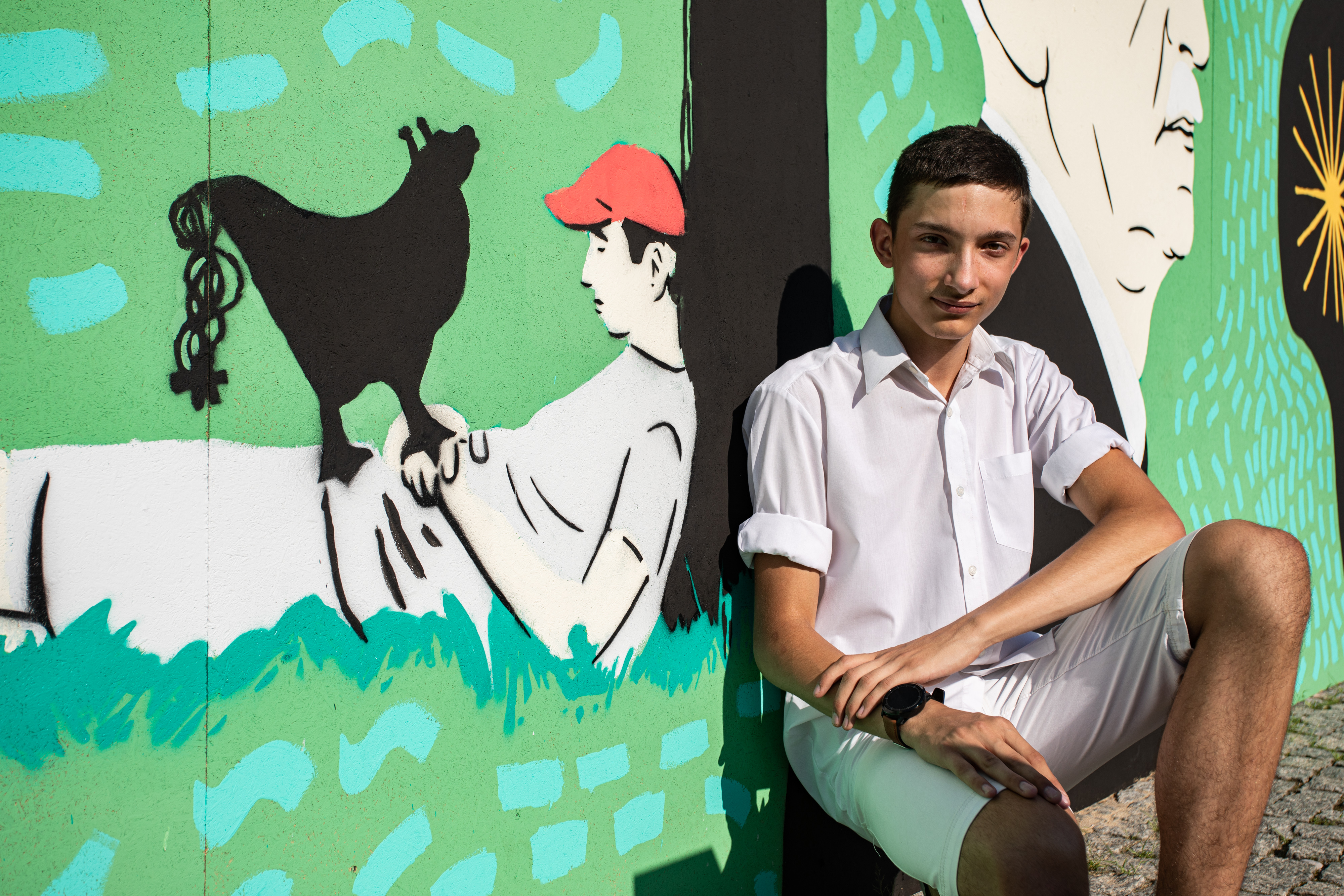 Uczestnik warsztatów na tle fragmentu muralu na Łące Leśmiana przedstawiający odpoczywającego chłopca oraz kota.