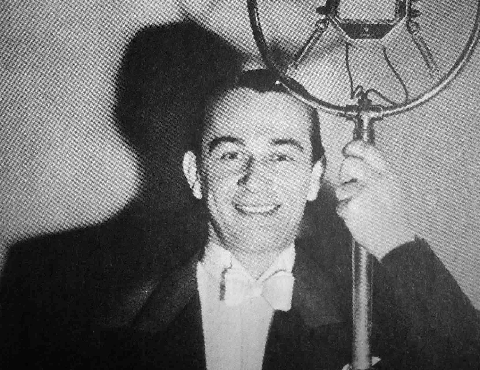Czarno-białe zdjęcie przedstawiauśmiechniętego mężczyznę (Mieczysław Fogg), ubrany we frak i białą koszulę z muszką, opiera się o starodawny mikrofon radiowy
