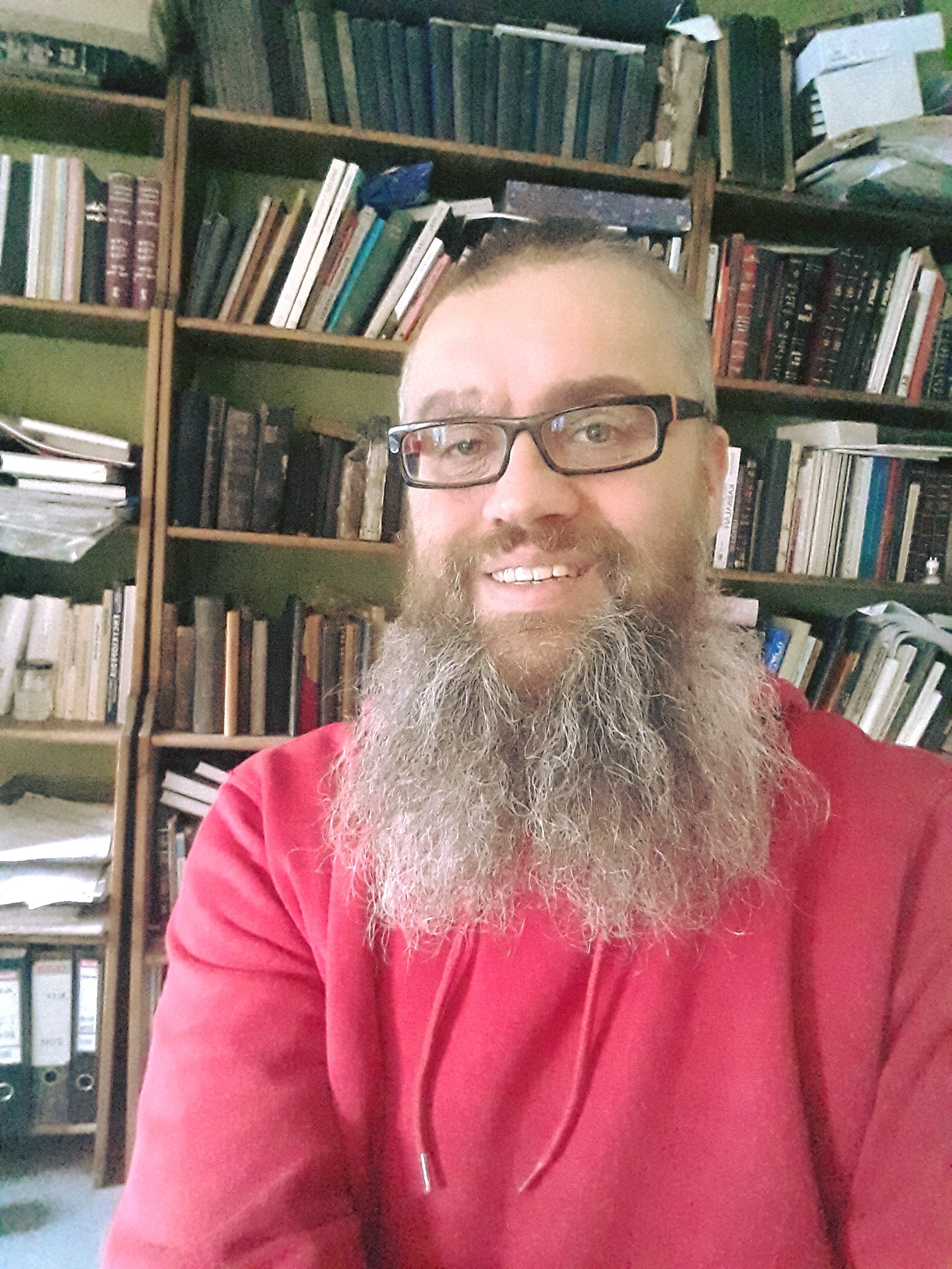 Portret mężczyzny - Dariusz Dekiert - okulary, długa broda, różowa sportowa bluza, w tle regały z książkami