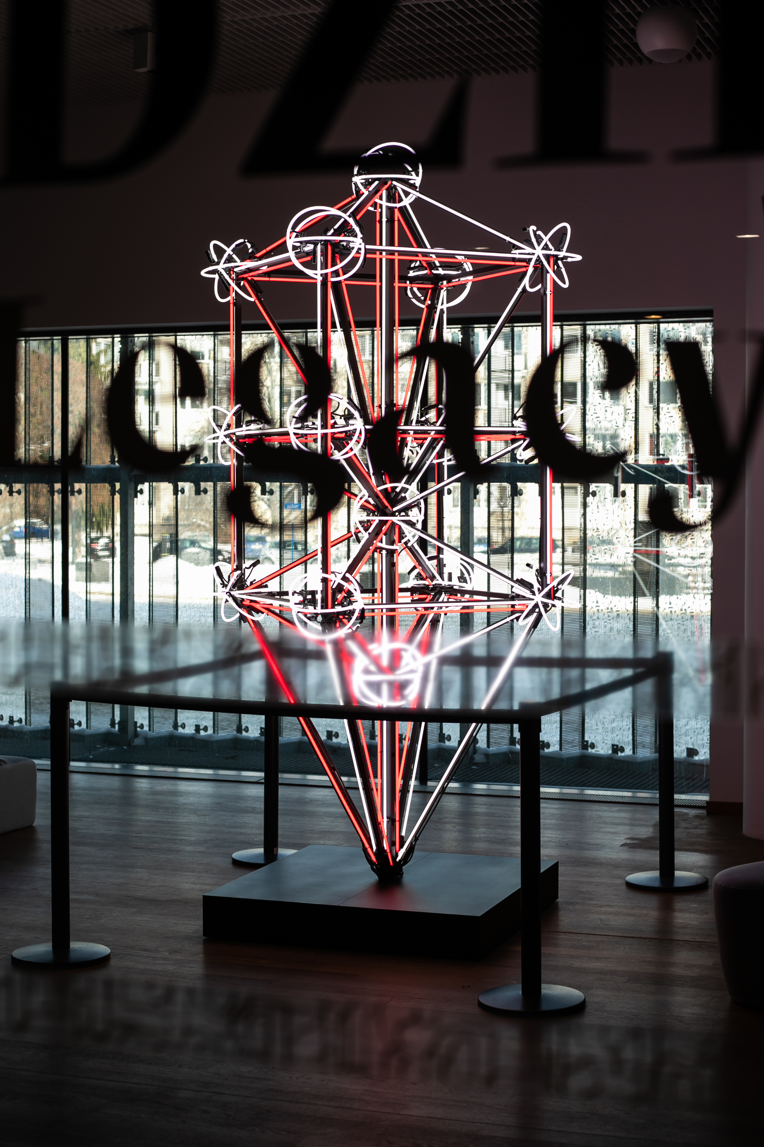Widok przez szybę z napisem Legacy na rzeźbę Huberta Czerepoka - z białych i czerwonych świetlówek spleciona bryła w kształcie kryształu