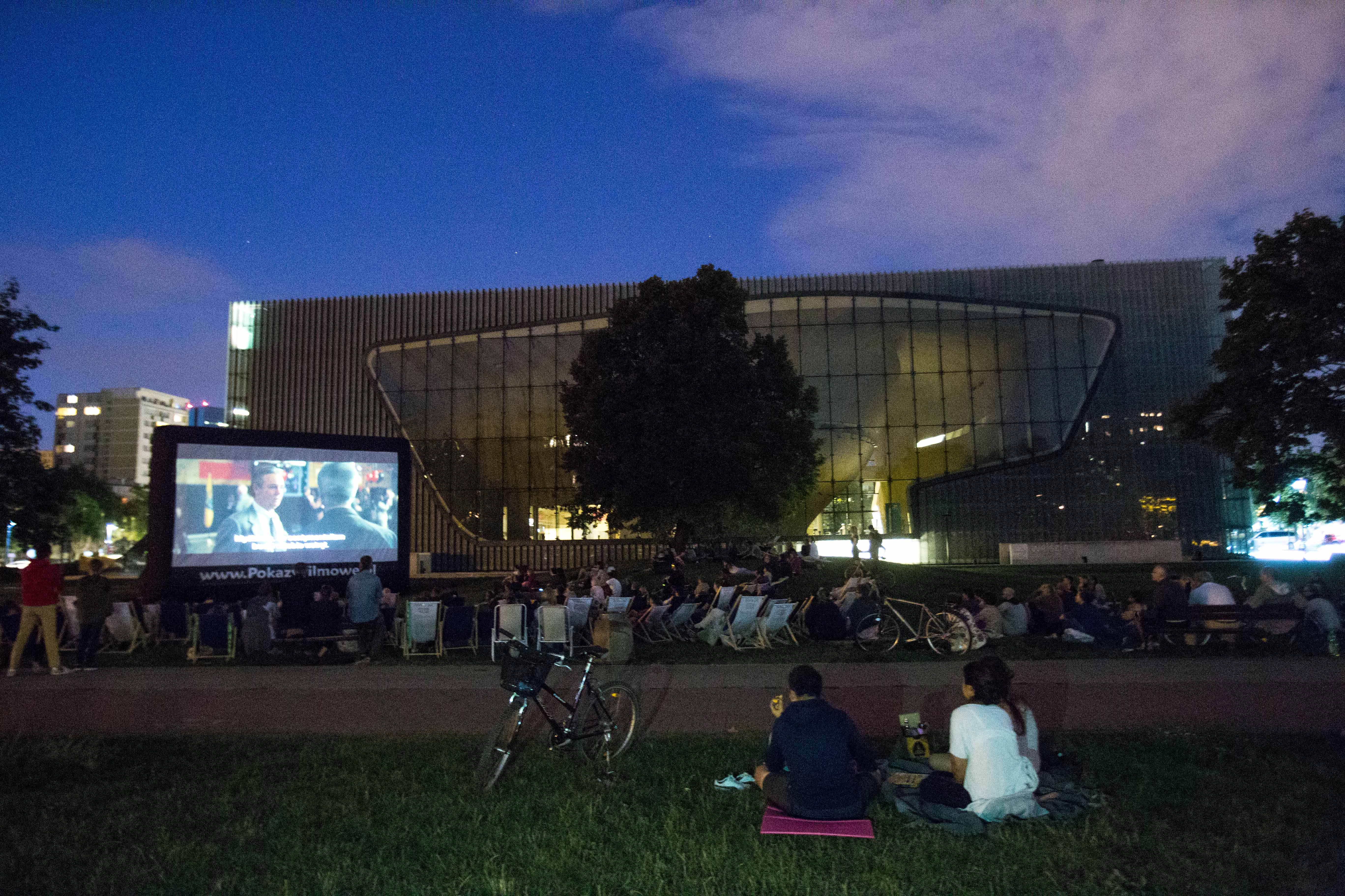 Na obrazie widzimy plenerowe kino letnie. Przed dużym ekranem znajduje się grupa widzów, oglądająca wieczorny seans film.