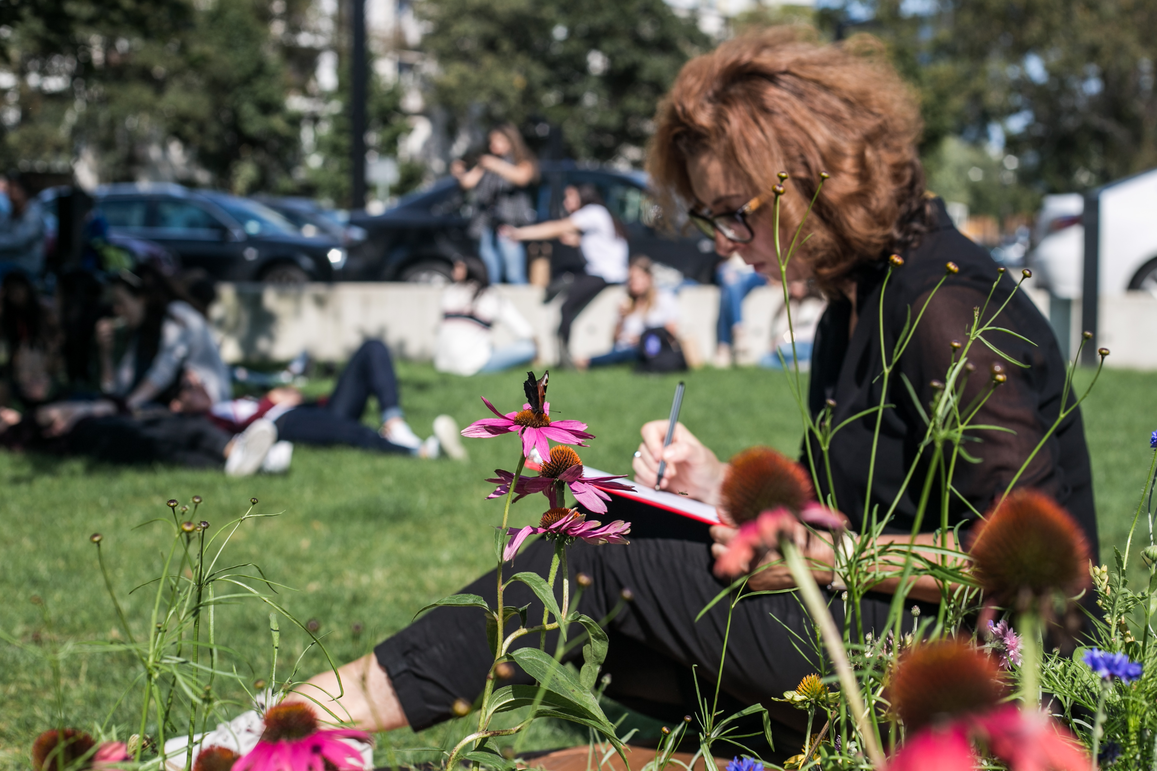 na obrazie widzimy kobietę siedząca na trawie, notująca w notesie opartym o kolana. 