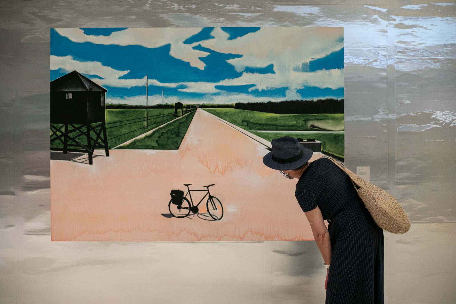 Osoba w czarnym ubraniu i czarnym kapeluszu pochyla się przed obrazem Wilhelma Sasnala, na którym przedstawiony jest rower, który stoi przed obozem zagłady w Bełżcu