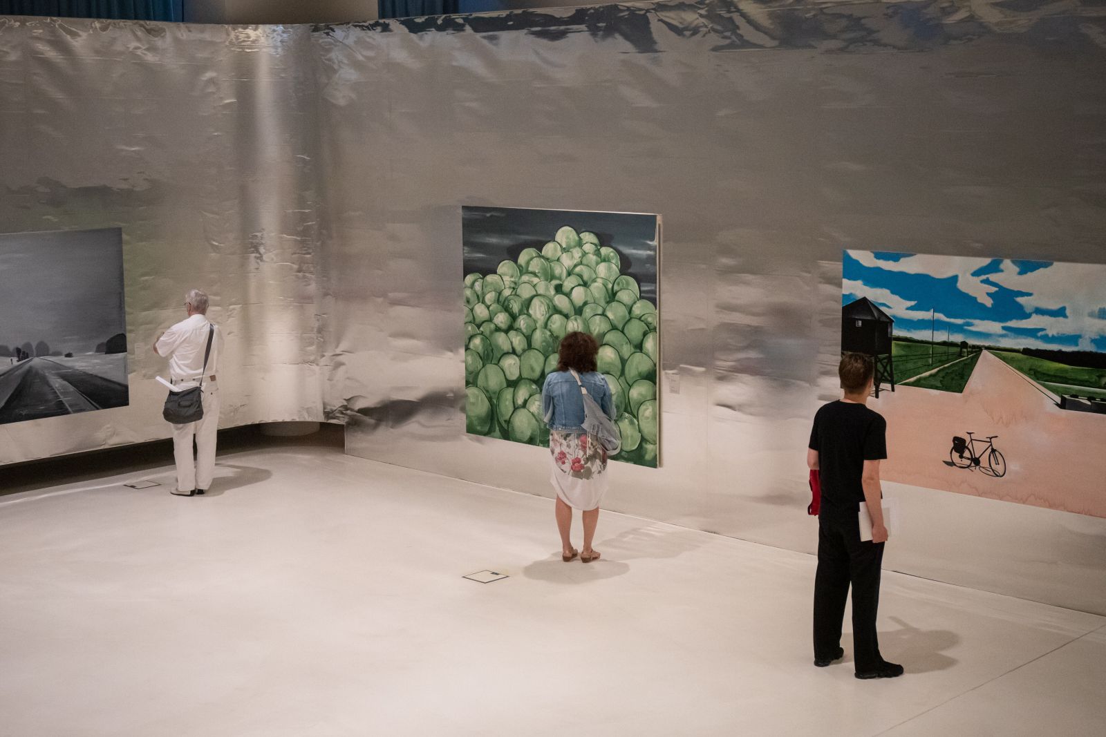 W przestrzeni wystawy Wilhelma Sasnala przed trzema obrazami wiszącymi obok siebie stoi troje ludzi