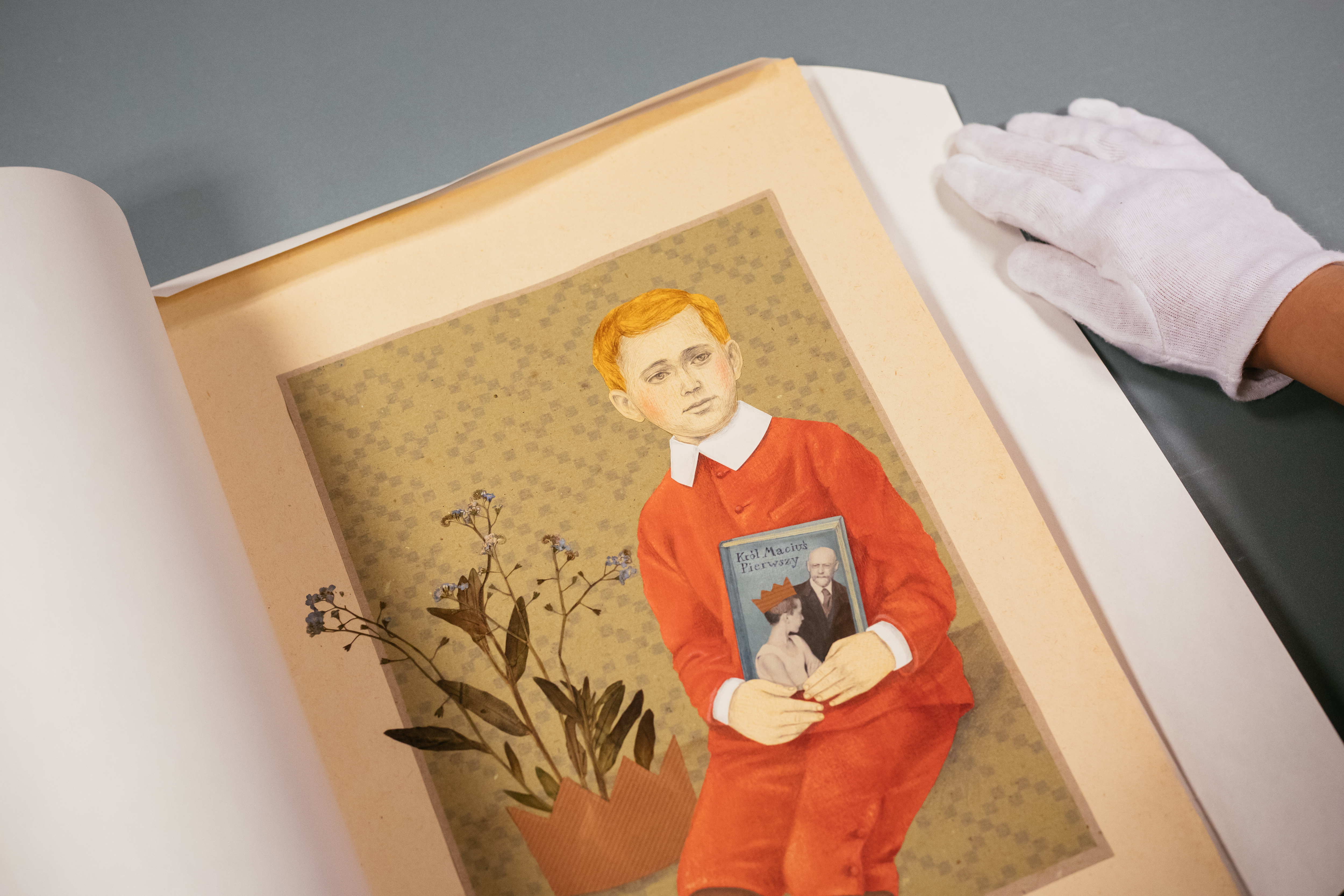 Obraz przedstawiający króla Maciusia -rudego chłopca w czerwonej marynarce i czerwonych spodniach do połowy nóg, trzymającego przy pasie, okładką do widza, książkę. Na lewo od chłopca - niebieskie kwiatki, wystające z brązowej schematycznej korony.