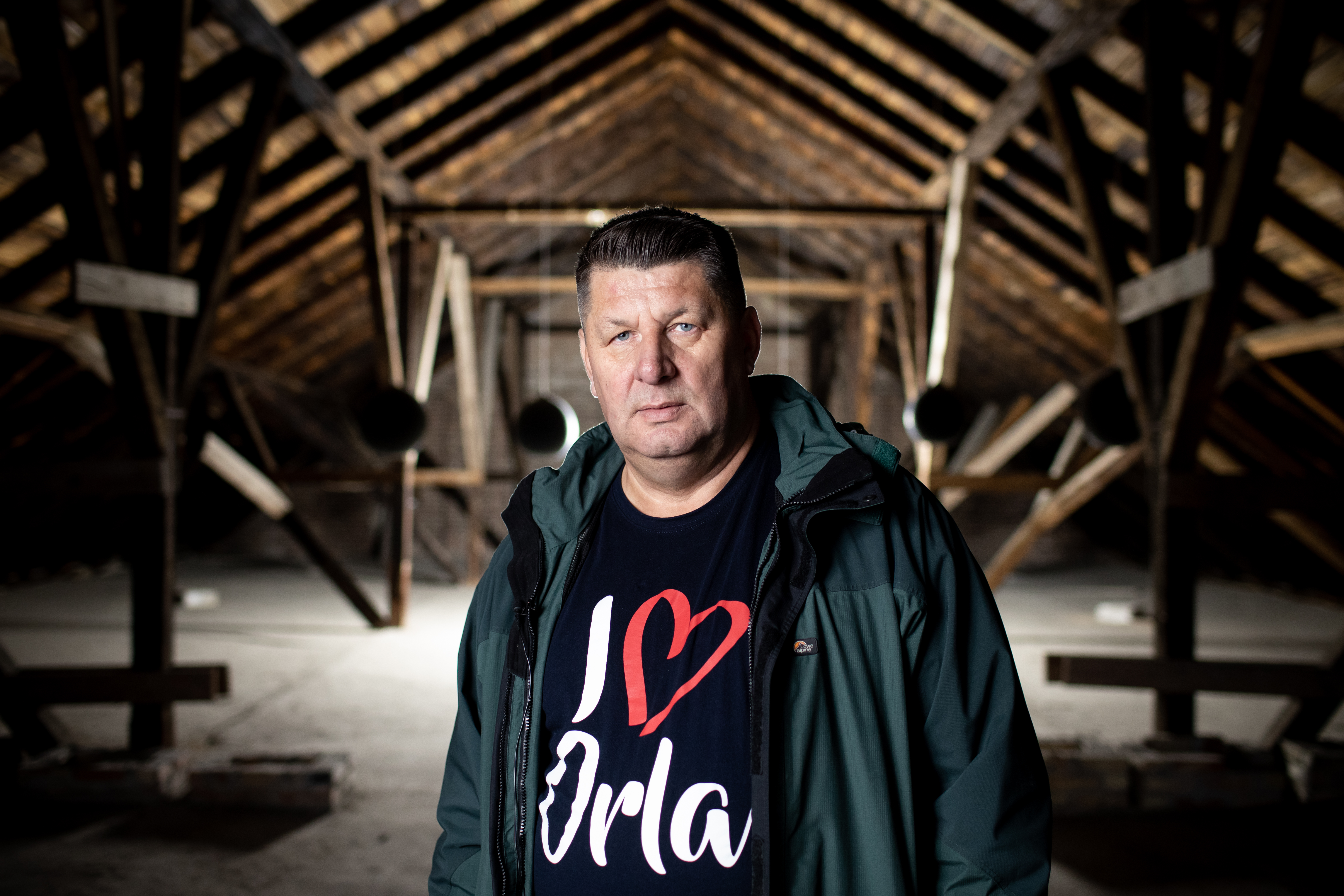 Marek Chmielewski stoi na poddszu drewnianego domu, widać żebrowanie dachu. Ubrany jest w czarną koszulkę z napisem "I <3 Orla"