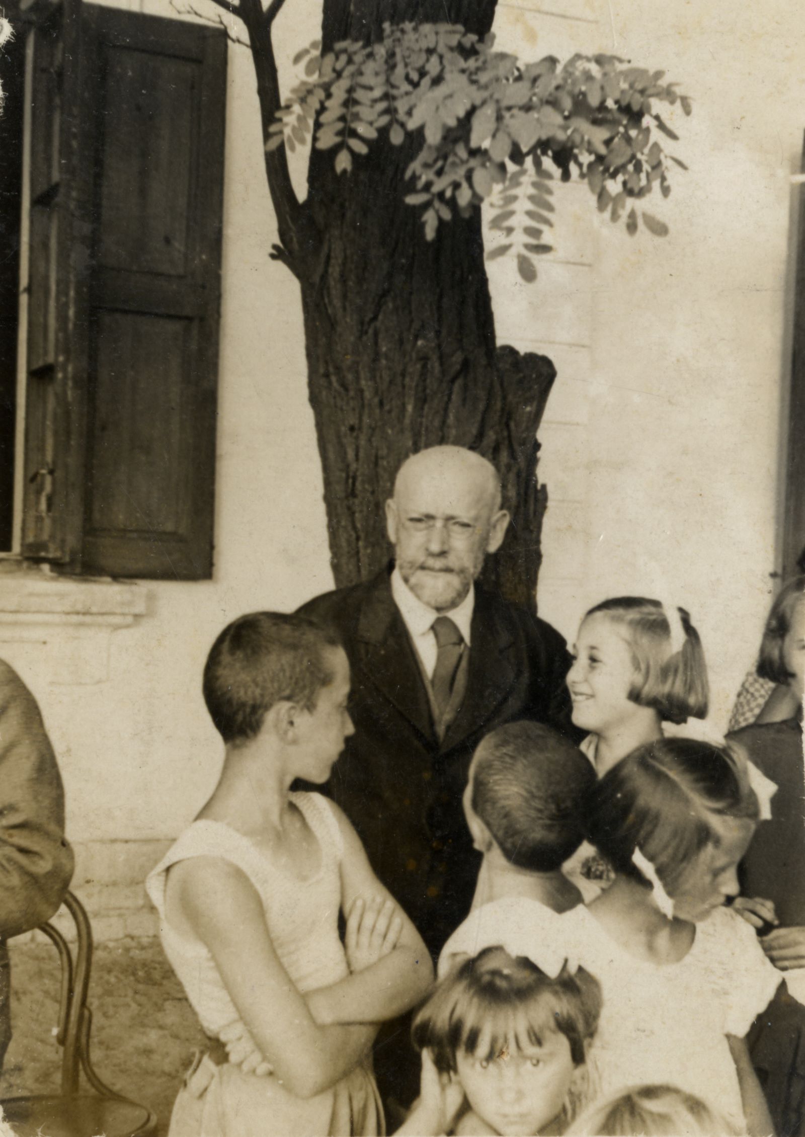 Czarno-białe zdjęcie. Janusz Korczak - łysy mężczyzna z wąsem i w okularach - w otoczeniu dzieci.