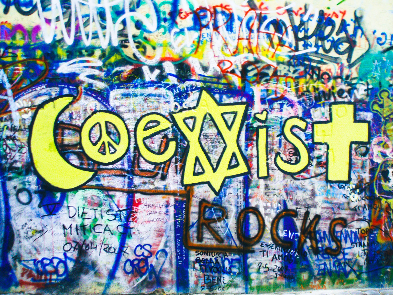 Ściana Johna Lennona w Pradze, na niej napis Coexist. X jest w kształcie Gwiazdy Dawida. Pod spodem dopisek rocks, czyli Coexist rocks.