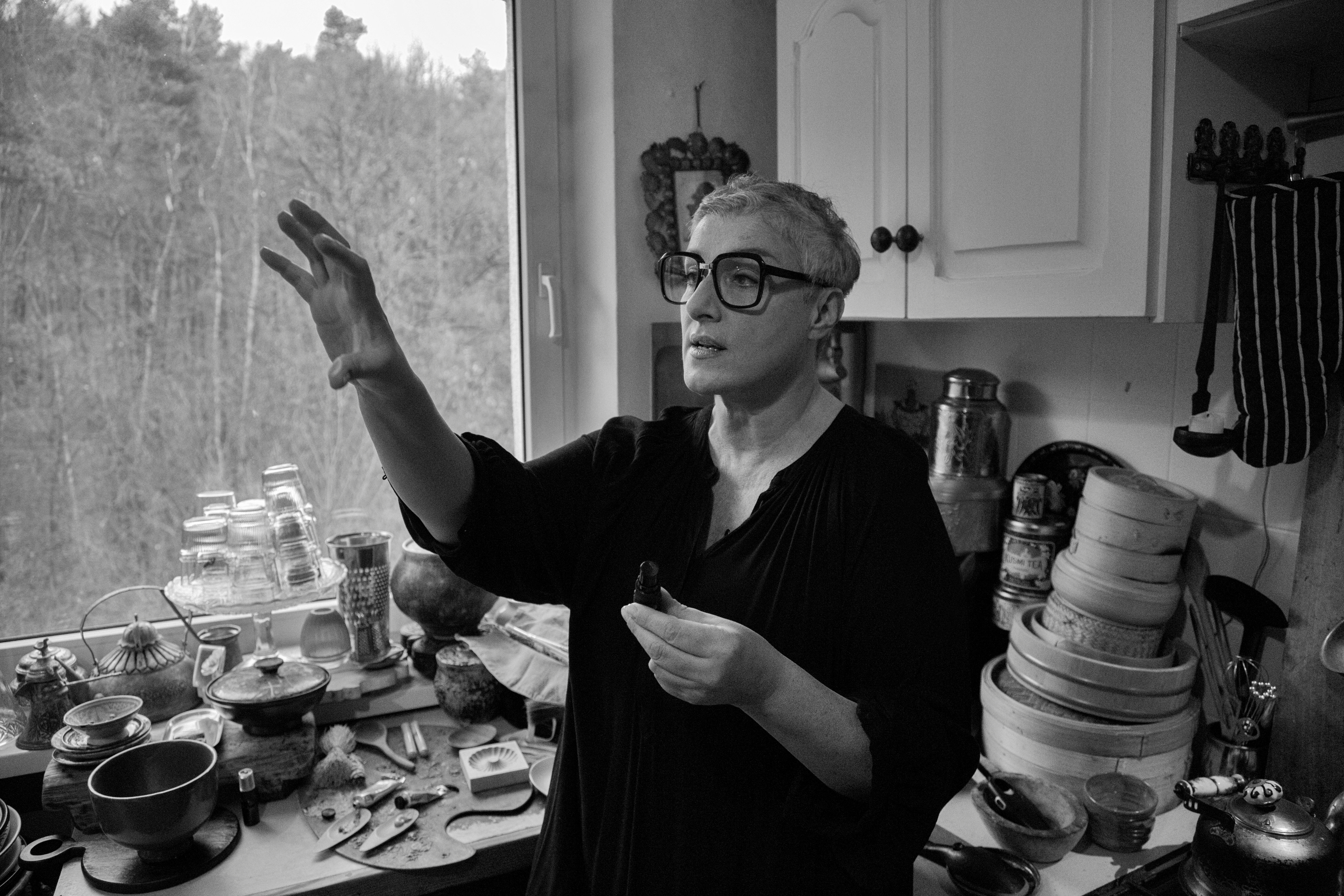 Czarno-białe zdjęcie przedstawia Annę Królikiewicz. Artystka ma okulary z okrągłymi szkłami, jedną rękę podnosi do góry.