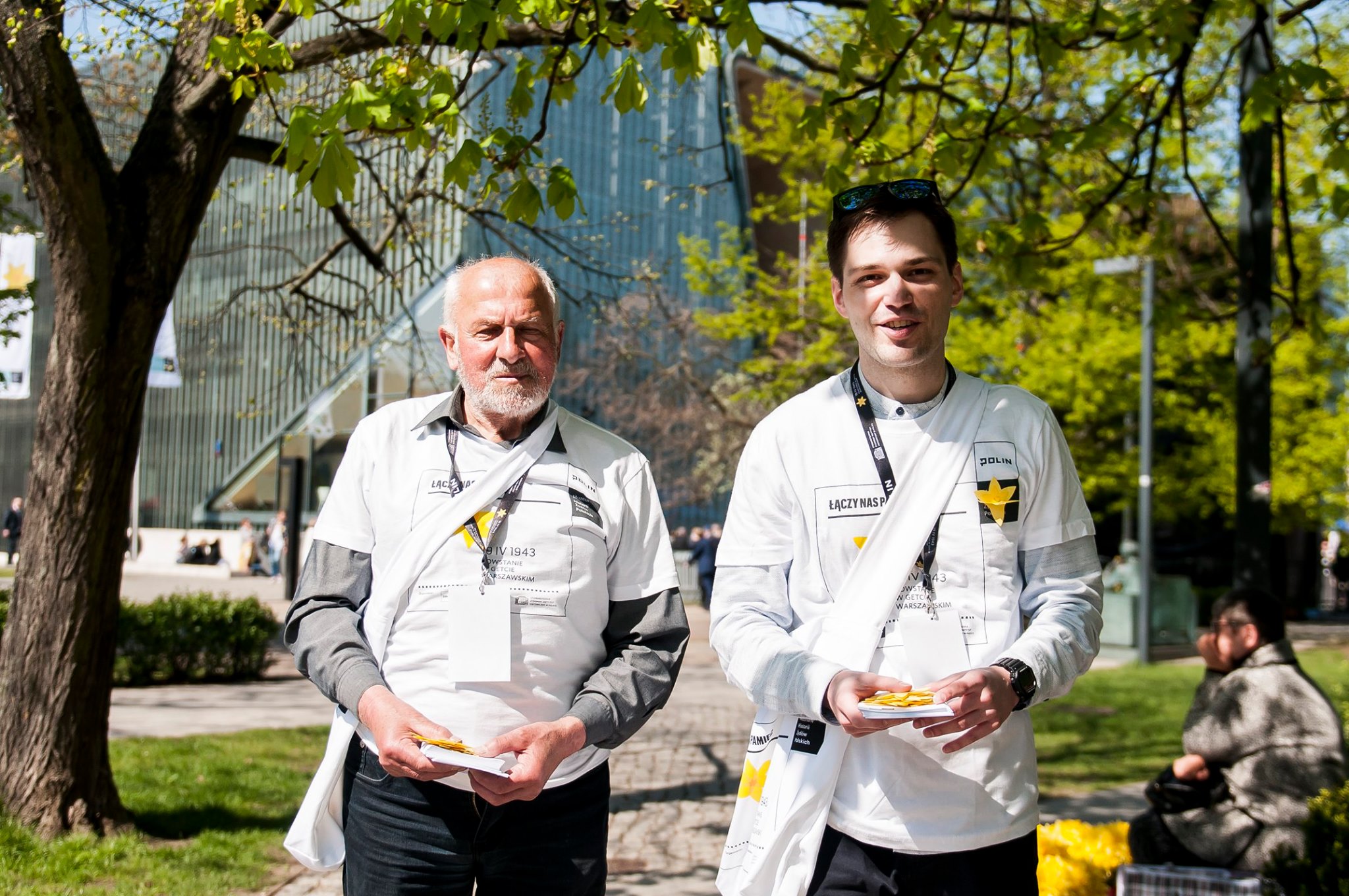 Dwaj (starszy i młody) wolontariusze w parku przed budynkiem Muzeum POLIN.