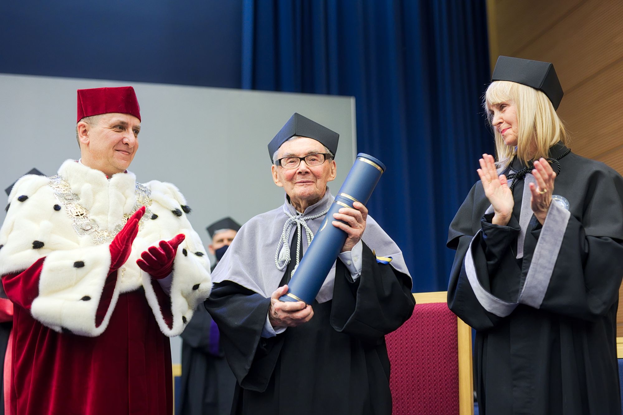 Marian Turski odebrał tytuł honoris causa. Trzyma tubę. Obok stoją rektor i promotorka nadania tytułu i biją brawo.
