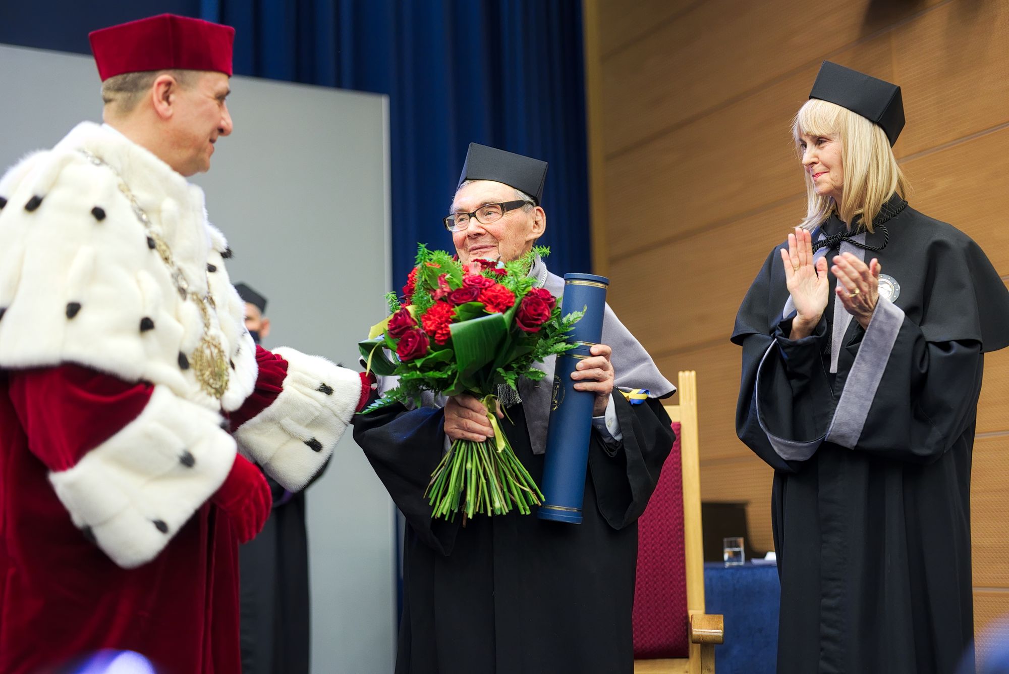 Marian Turski trzyma kwiaty i tubę, w której znajduje się dokument potwierdzający tytuł honoris causa. Patrzy w stronę rektora UMCS. Obok brawo bije promotorka nadania tytułu.