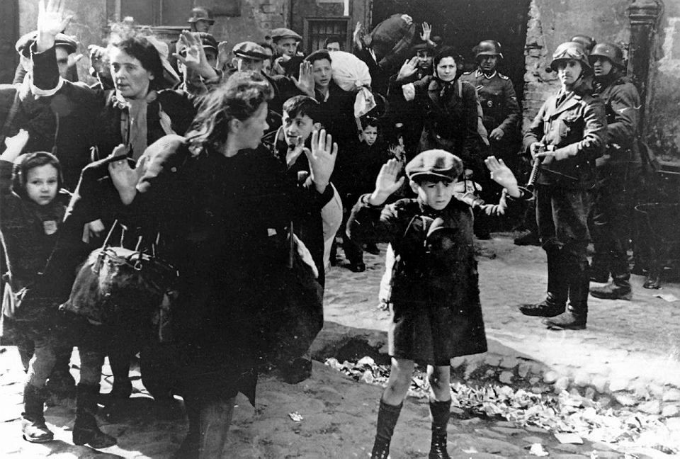 Czarno-biała fotografia przedstawia Żydów wyciągniętych z bunkrów. Podnoszą ręce do góry w geście poddania się. Za nimi stoją mężczyźni w hełmach i z bronią. 