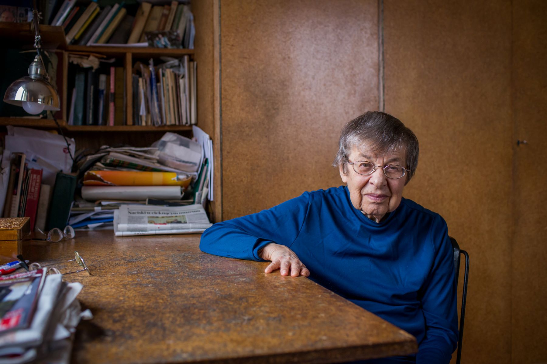 Barbara Góra - starsza kobieta w okularach - siedzi przy stole. Za jej plecami regał pełen książek.