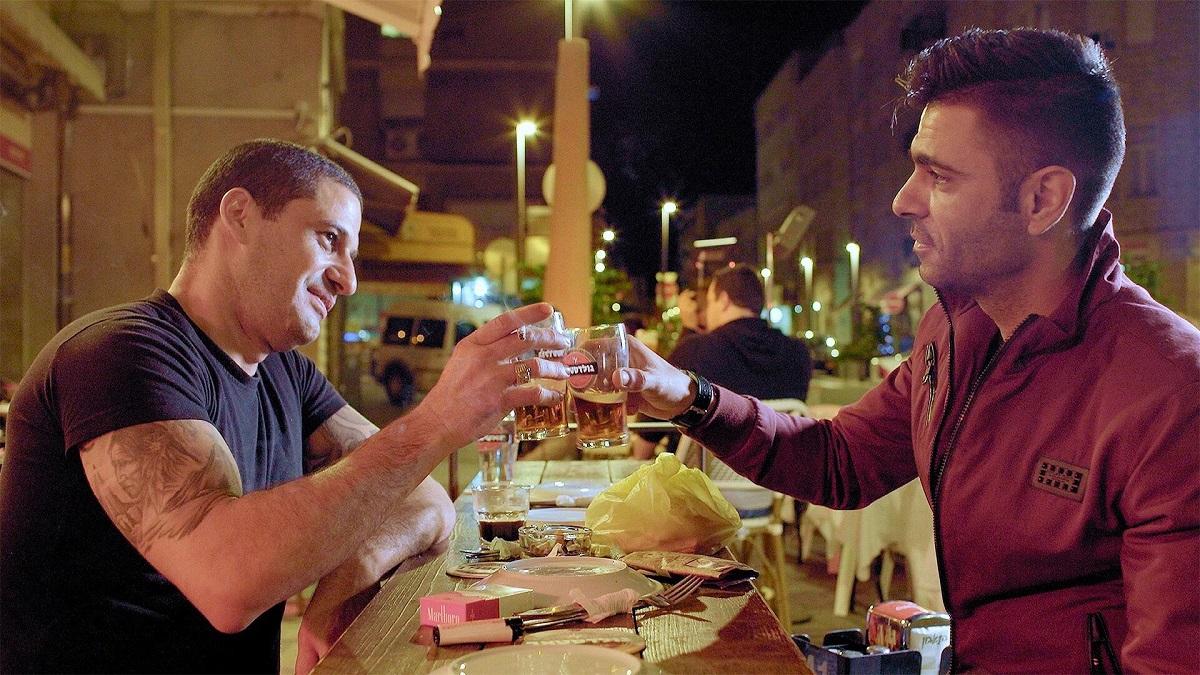 Kadr z filmu "Breaking Bread". Dwaj mężczyźni siedzą przy stole i stukają się szklankami z piwem.