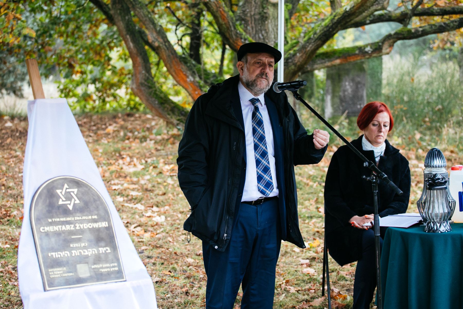 Naczelny Rabin Polski Michael Schudrich mówi przez mikrofon. Stoi obok tablicy upamiętniającej cmentarz żydowski w Górze Kalwarii. Obok przy stoliku siedzi kobieta.