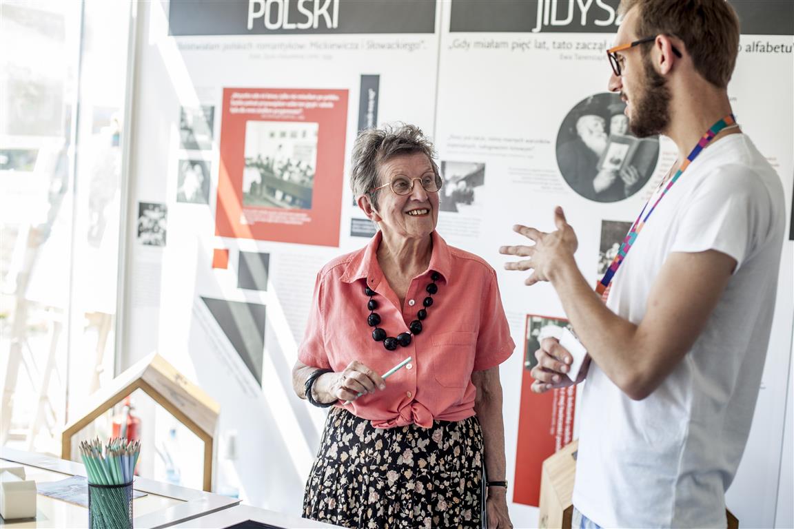 Starsza kobieta i młody mężczyzna rozmawiają ze sobą podczas spotkania na wystawie "Muzeum na kółkach". Za nimi, w tle banery z napisami polski i jidysz.