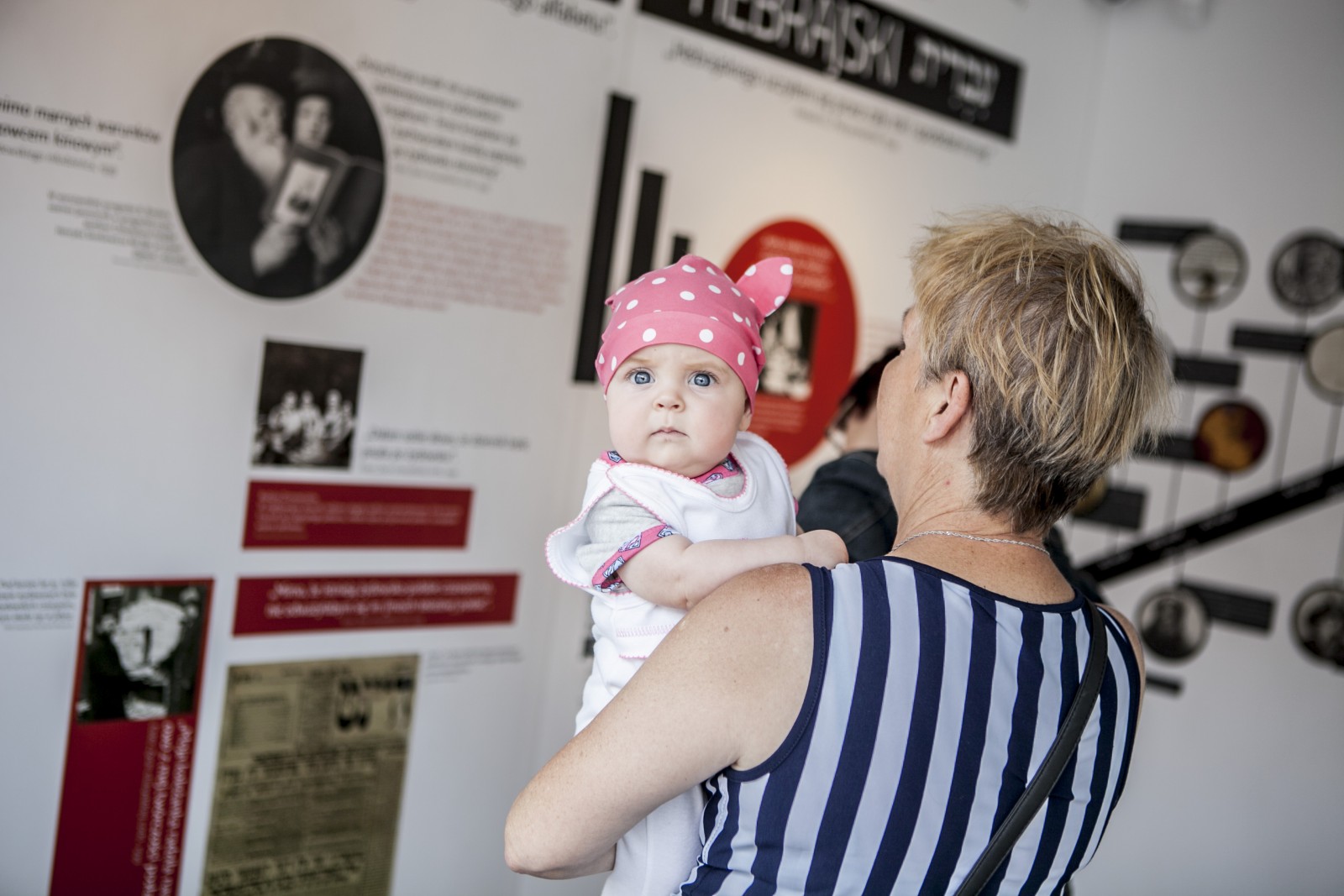 Starsza kobieta ogląda wystawę "Muzeum na kółkach" prezentowaną w Łazach. Trzyma na rękach niemowlę.