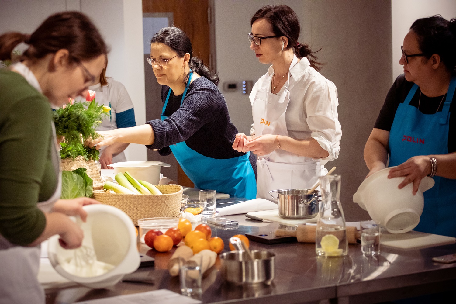 Kilka kobiet w fartuchach stoi przy stole podczas warsztatów kulinarnych.