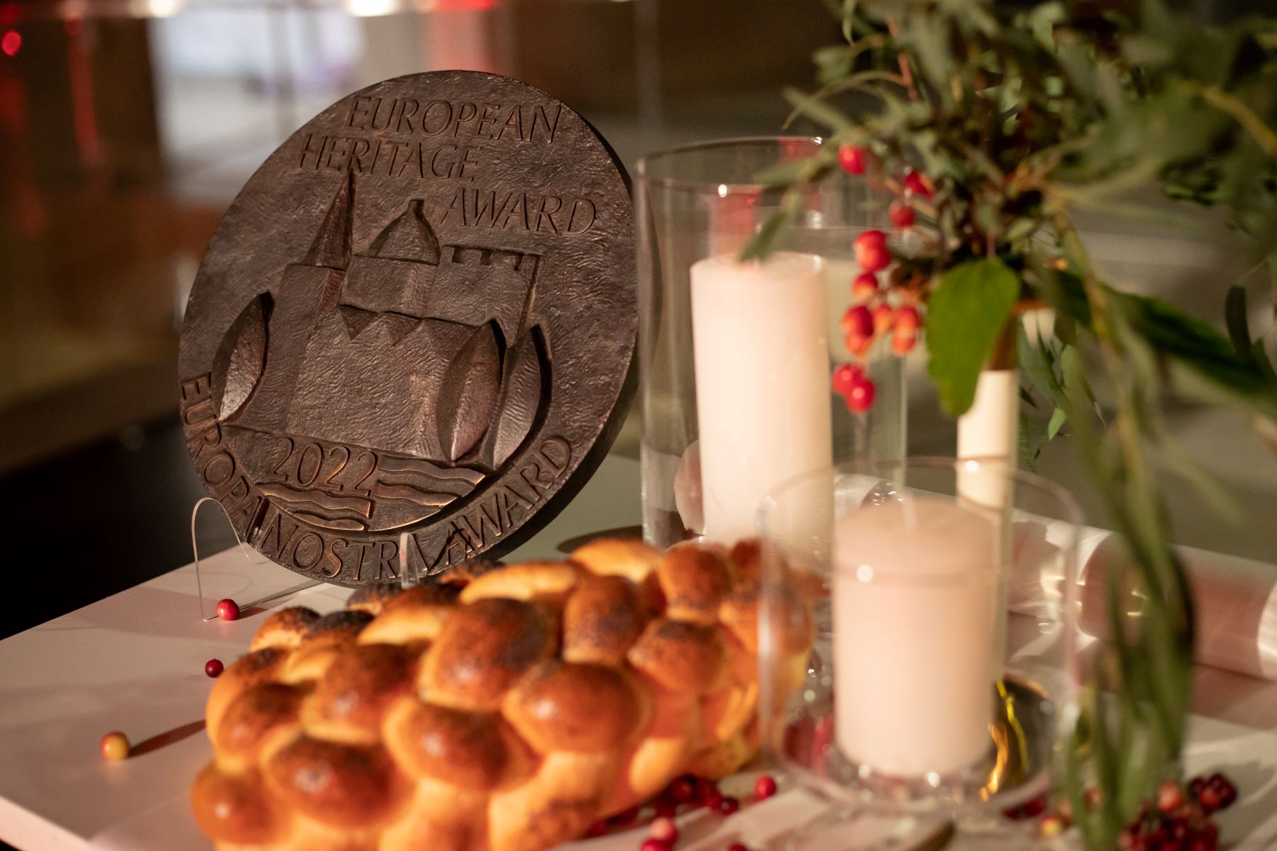 Na stole przy chałce, roślinnej dekoracji i świeczkach stoi medal Nagrody Europa Nostra 2022.