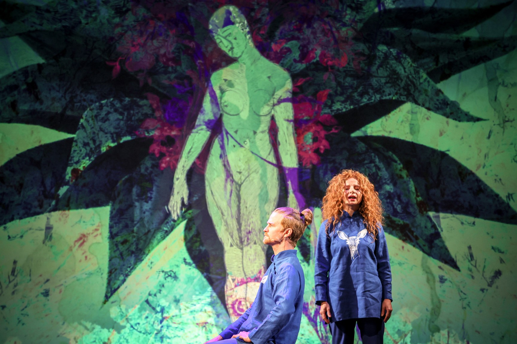 Kadr ze spektaklu "Natura ludzka". Kobieta stoi, mężczyzna siedzi. Oboje mają rude włosy. Za nimi grafika przedstawiająca człowieka na tle ciemnego rozchylonego kwiatka.
