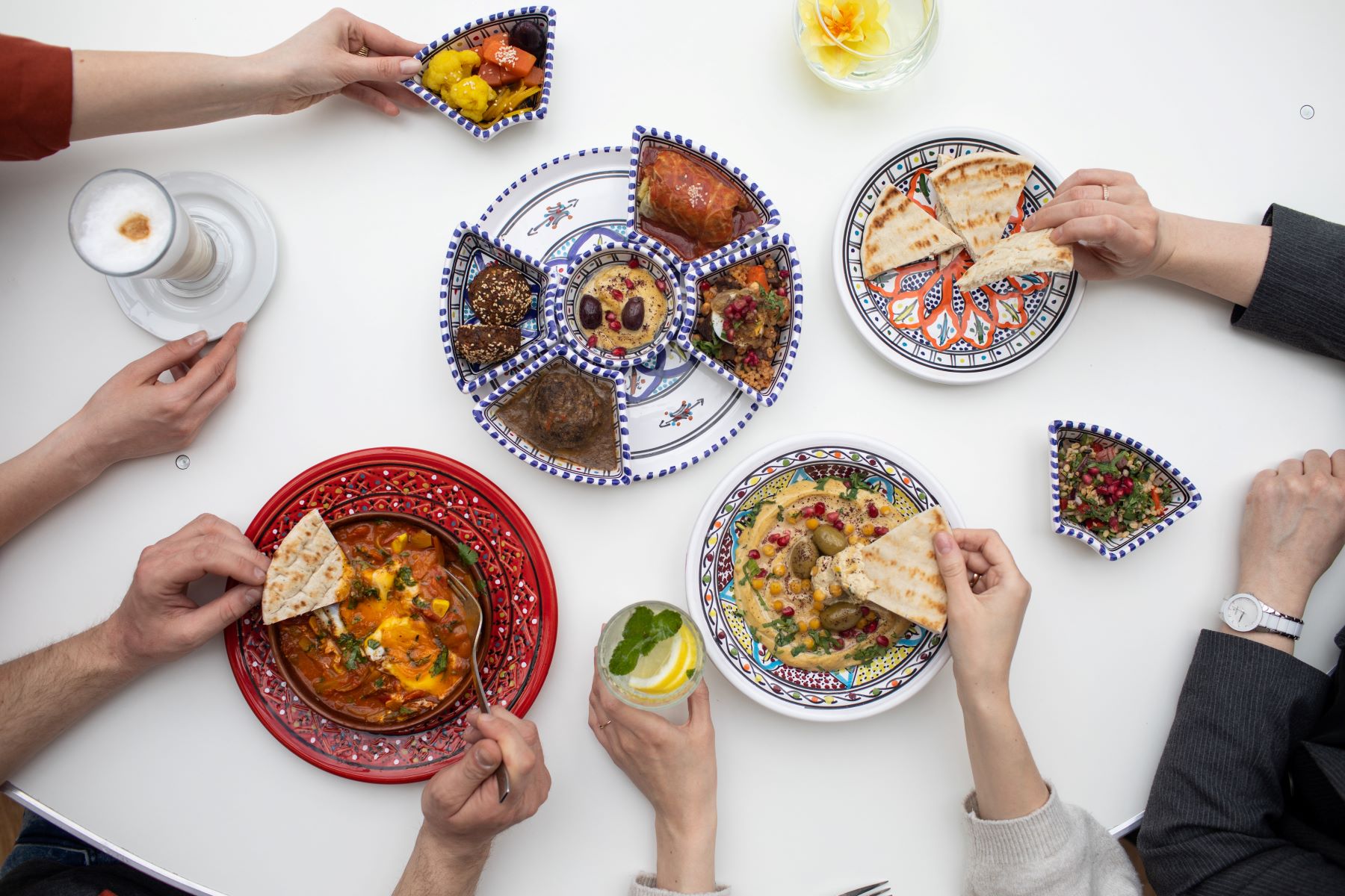 Na stole w miskach stoją potrawy: pita, szakszuka, humus, gołąbki, sałatka izraelska. Kilka osób degustuje potrawy.