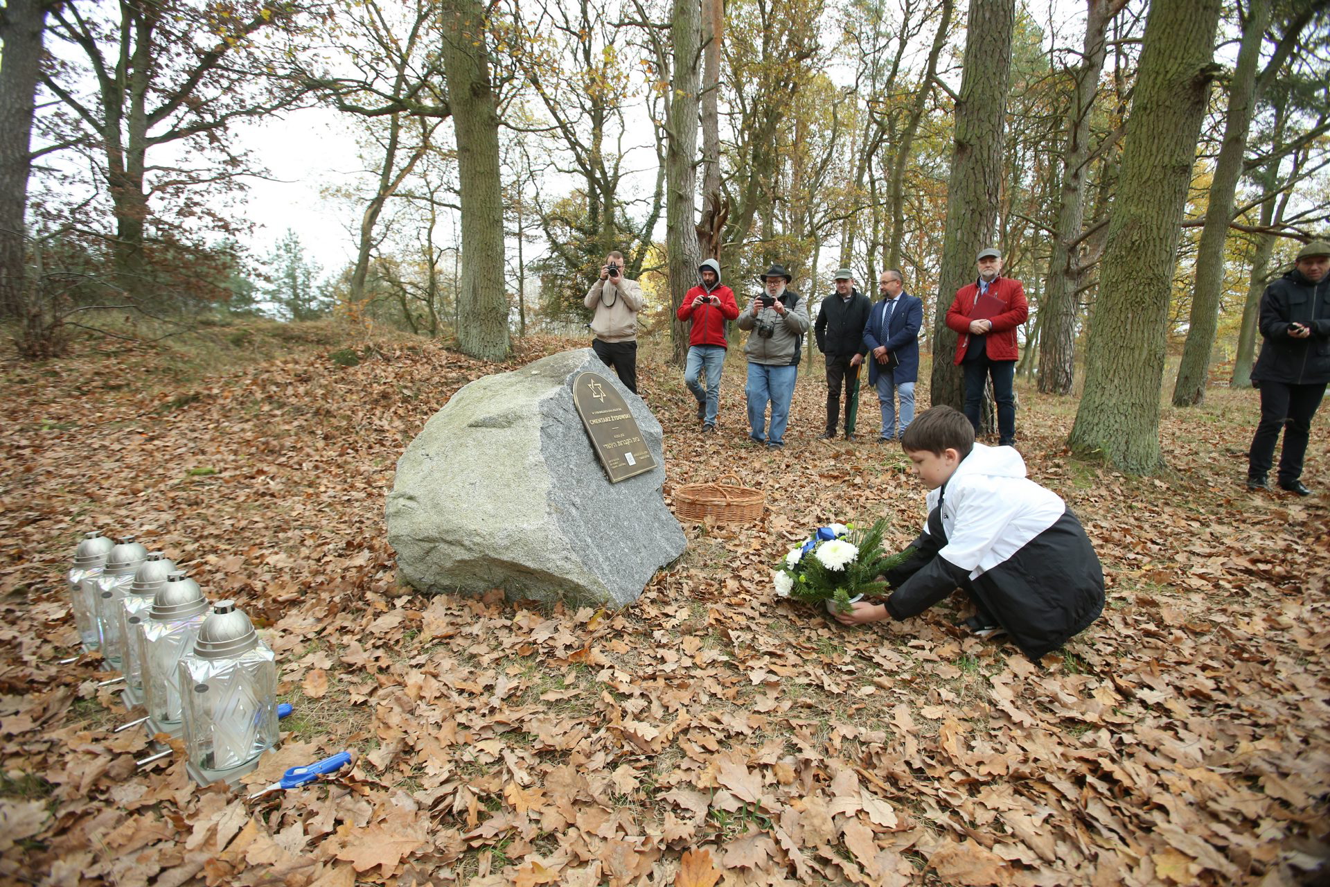 Chłopiec kładzie kwiaty pod kamieniem z tablicą upamiętniającą cmentarz żydowski w Stepnicy. Kilka osób robi mu zdjęcie.