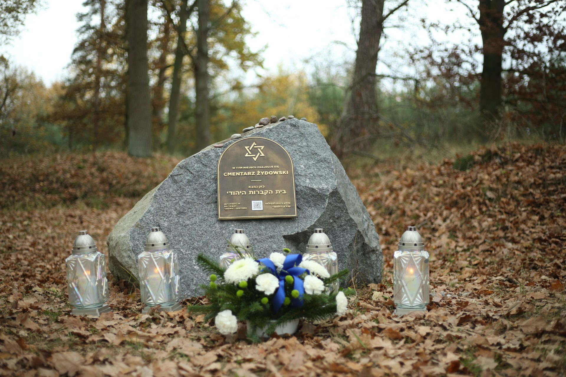 Pod tablicą upamiętniającą cmentarz żydowski w Stepnicy stoją zapalone znicze i leży mała wiązanka z kwiatów.