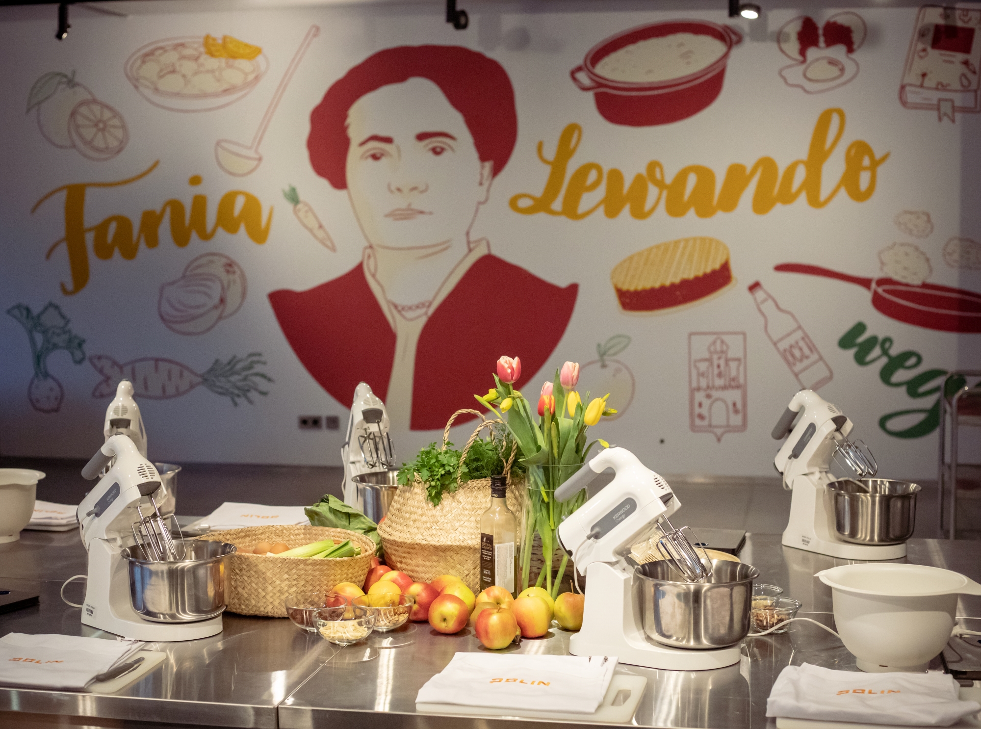 Na stole w pracowni kulinarnej "u Fani" leżą jabłka, stoją garnki i miksery oraz kwiaty w wazonie. W tle mural z podobizną Fani Lewando.