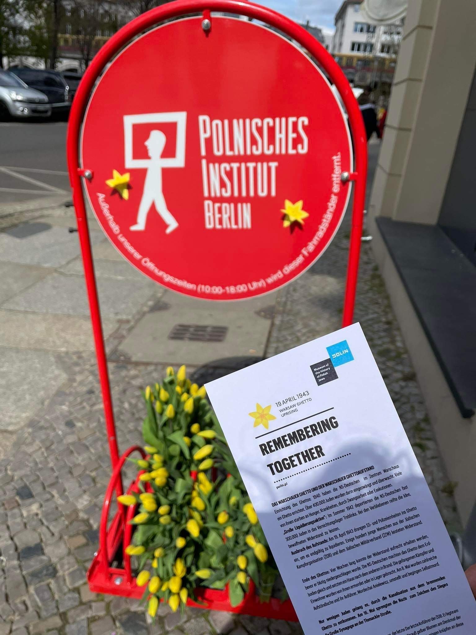 Red sign of Polish Institute in Berlin with daffodils on it. Next to it English leaflet of Warsaw Ghetto Uprising Campaign.