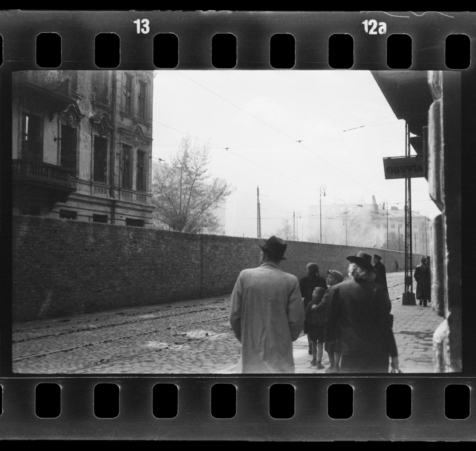 Kilka spacerowiczów na placu Muranowskim. Spoglądają w lewo w stronę muru i budynku na terenie getta warszawskiego.