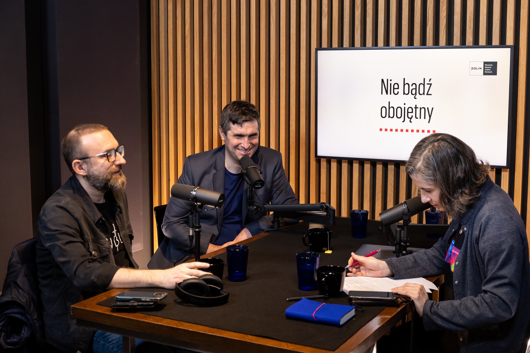 W studiu, podczas nagrania pierwszego odcinka podcastu siedzą przy stole: Karolina Wigura, Tomasz Stawiszyński i Piotr Stankiewicz. W tle na ekranie napis Nie bądź obojętny.