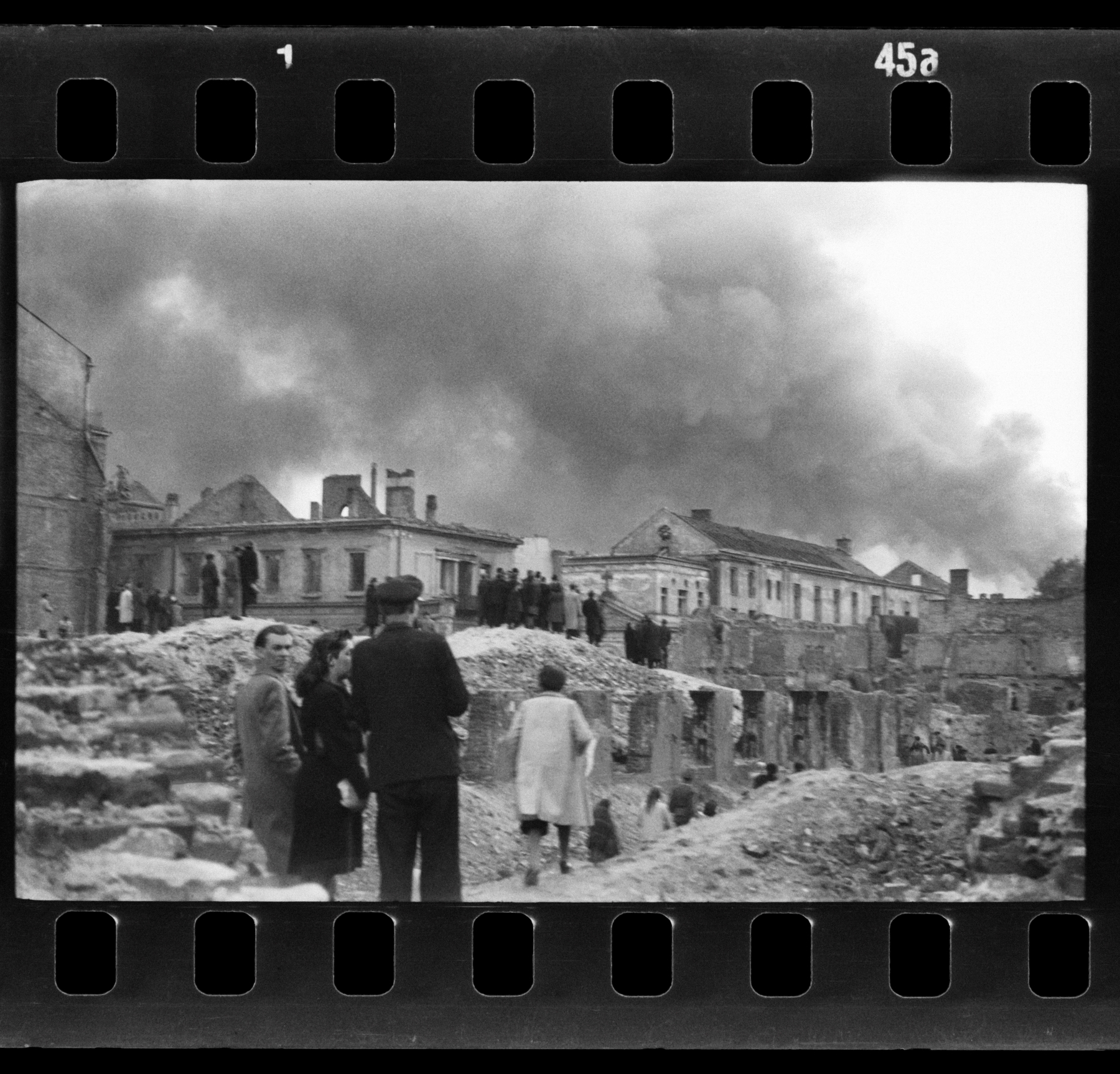 Kilka osób stoi przy zniszczonych zabudowaniach szpitala św. Ducha. W tle dym płonącego getta.