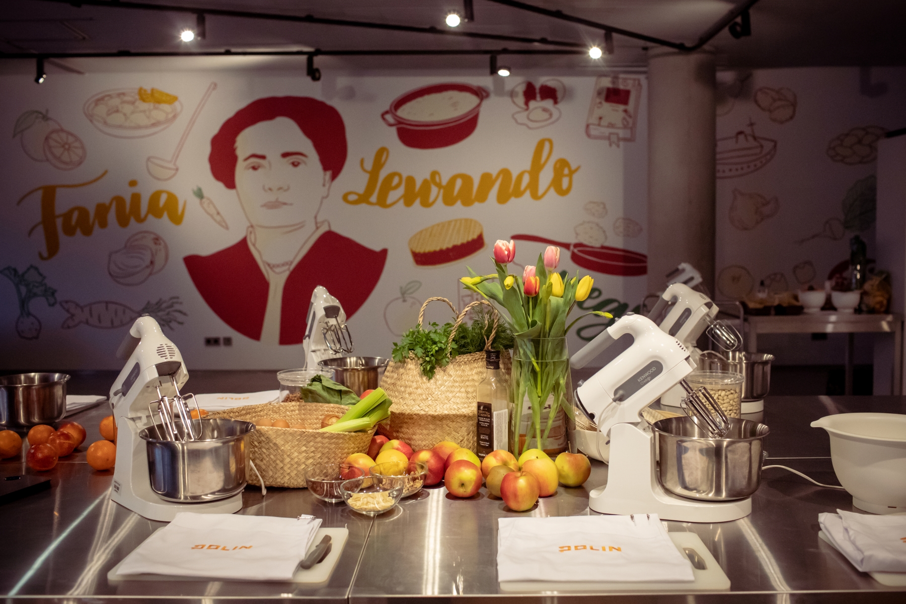 Na stole w Pracowni kulinarnej "U Fani" stoją miksery, wazon z kwiatami, leżą jabłka, mandarynki i serwetki. W tle mural z Fanią Lewando.