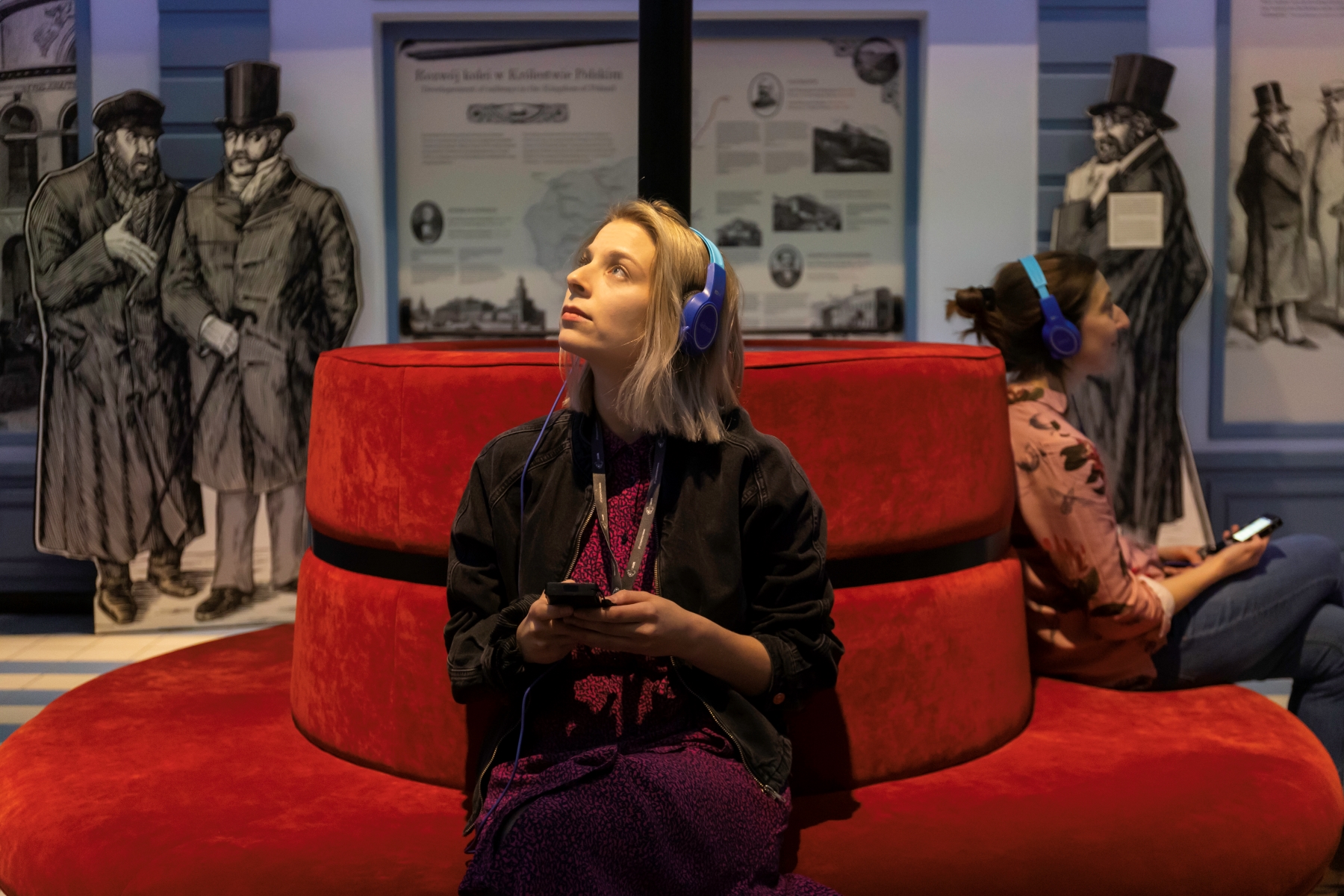 Dwie kobiety siedzą na kanapie w jednej z galerii na wystawie stałej w POLIN. Mają słuchawki na uszach i słuchają nagrań z audioprzewodnika.