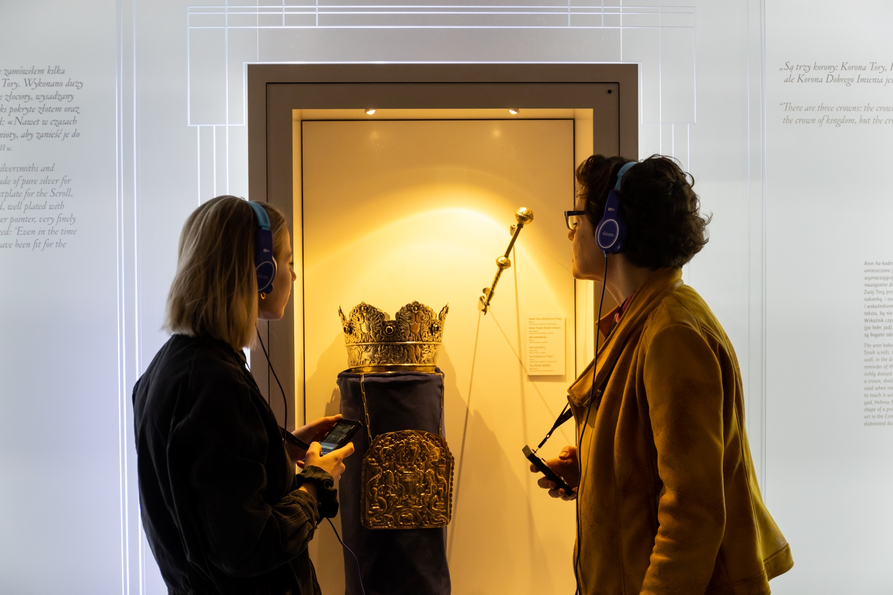 Dwie kobiety ze słuchawkami na uszach przyglądają się eksponatom na wystawie stałej.