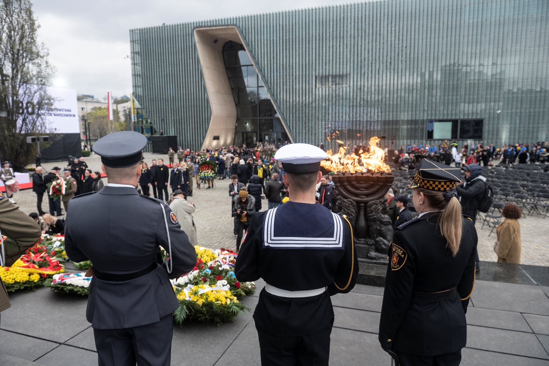 Straż złożona z trzech osób w mundurach stoi przy pomniku Bohaterów Getta. Jest zwrócona w kierunku budynku Muzeum POLIN. Do pomnika wędrują delegacje z kwiatami.