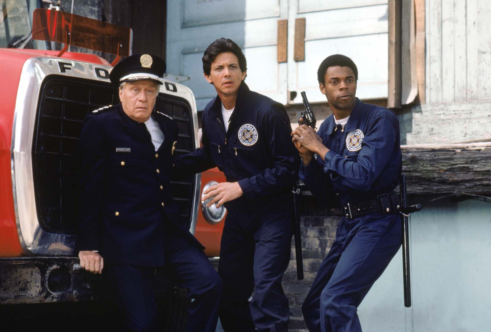 Kadr z filmu "Akademia policyjna". Trzej policjanci - jeden z nich, starszy, siedzi na samochodzie marki Ford, pozostali stoją obok niego.