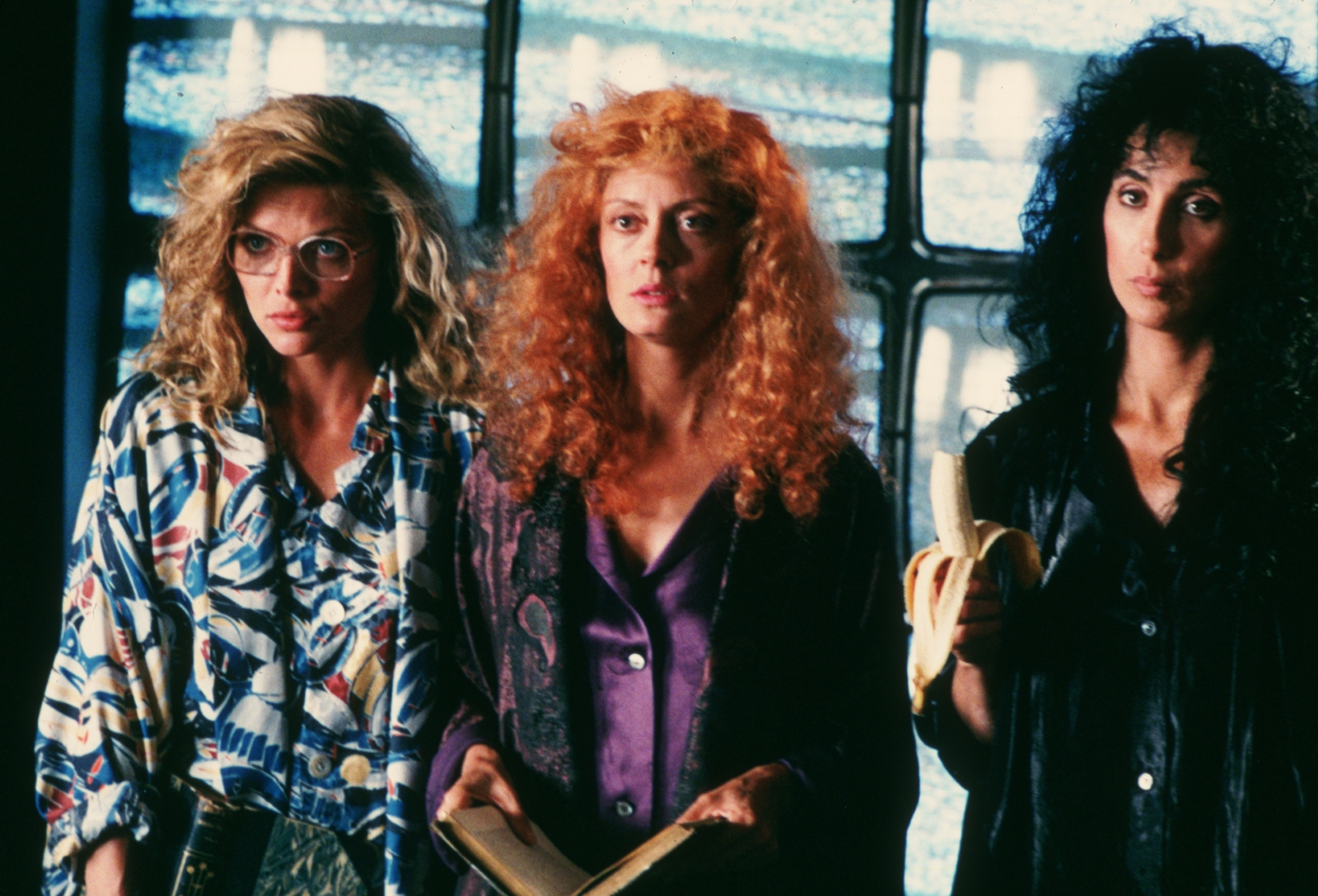 Kadr z filmu "Czarownice z Eastwick". Obok siebie stoją trzy kobiety o blond, rudych i czarnych włosach.
