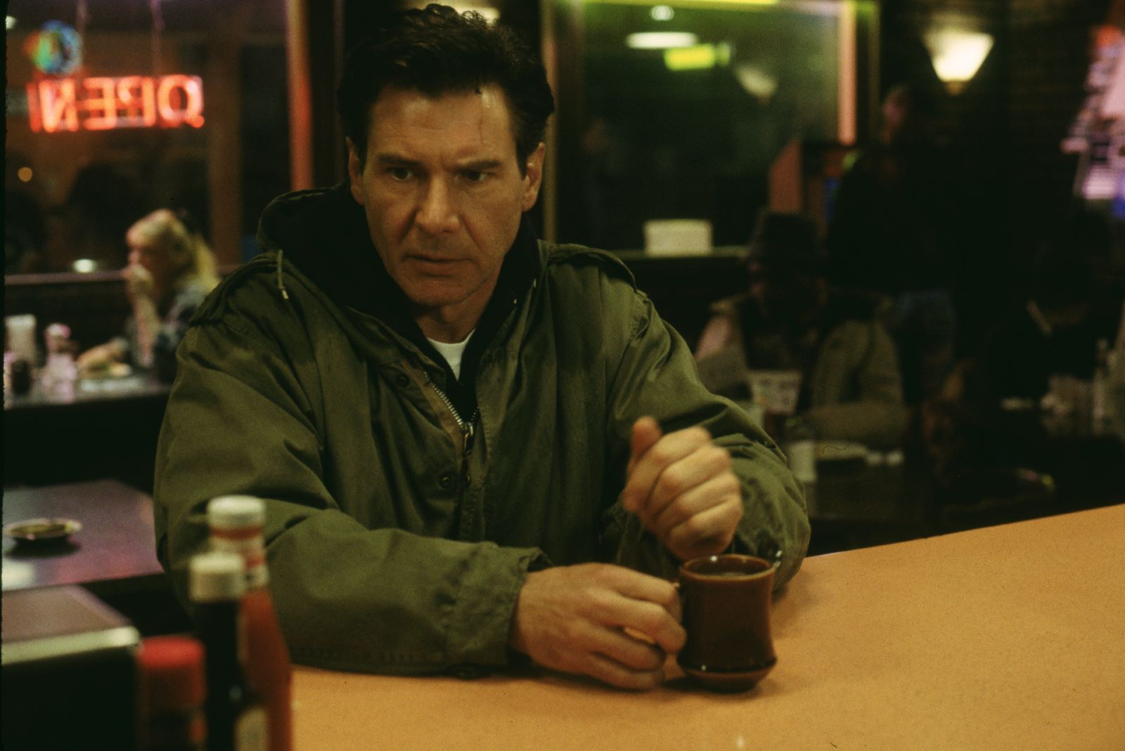 Kadr z filmu "Ścigany". Mężczyzna (Harisson Ford) siedzi przy stole i trzyma kubek.