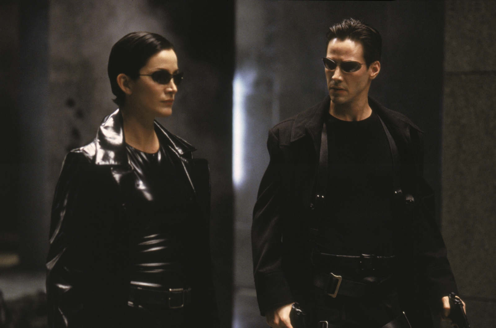 Kadr z filmu "Matrix". Obok siebie stoją kobieta i mężczyzna (główny bohater - Neo) w czarnych strojach i okularach.