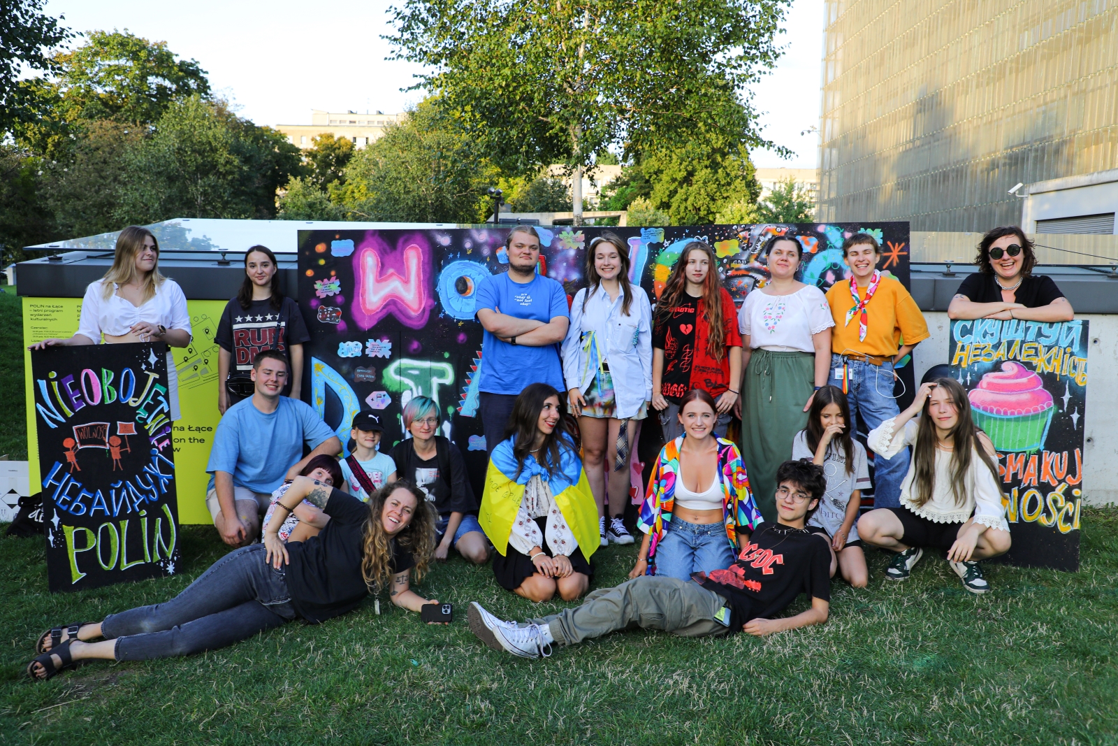 Grupa uczestników i uczestniczek warsztatów dla młodzieży siedzi lub stoi na trawie przy Muzeum POLIN.