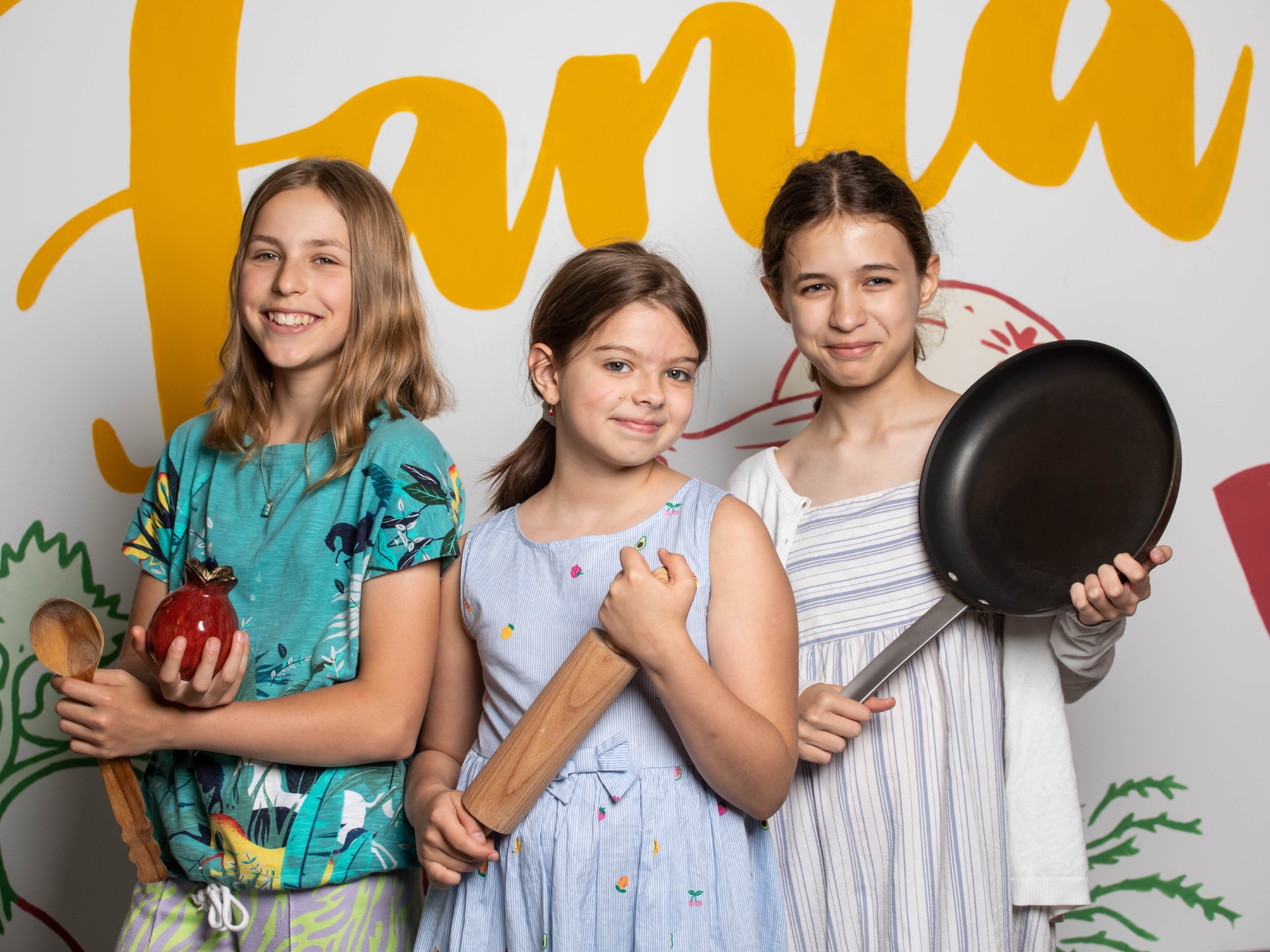 Trzy dziewczynki stoją obok siebie na tle muralu w Pracowni kulinarnej "U Fani". Każda z nich trzyma narzędzia kuchenne - łyżkę, wałek, patelnię.