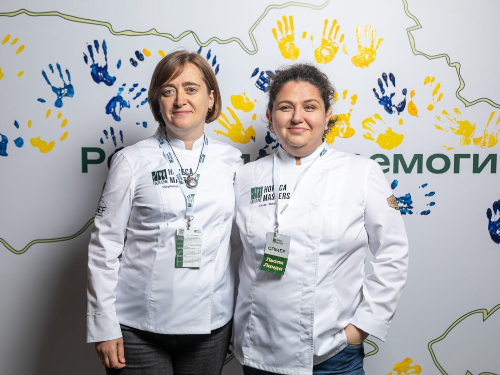 Dwie kobiety - Marianna Dushar i Lola Landa stoją obok siebie. W tle zarys terytorium Ukrainy, na którym odbite są dłonie w niebiesko-żółtych kolorach.