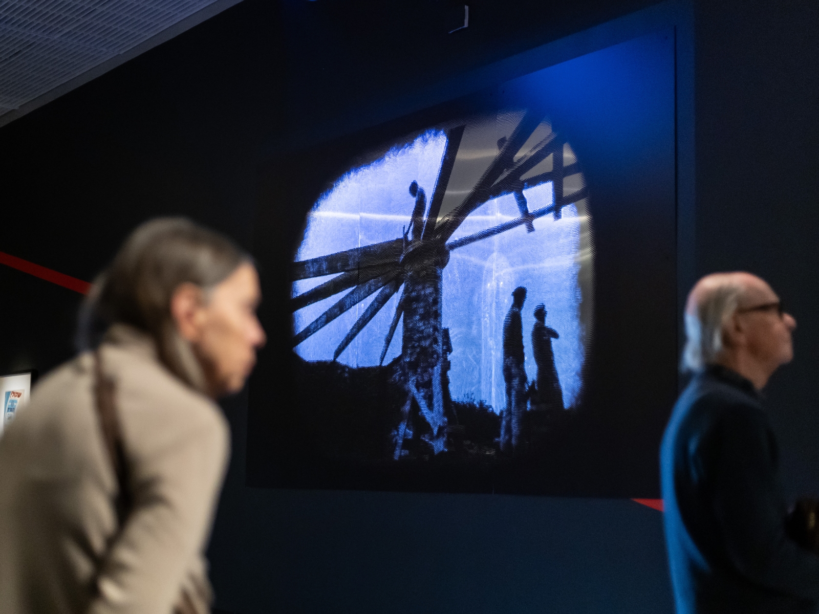 Kobieta i mężczyzna zwiedzają wystawę "1945. Nie koniec, nie początek". W tle podświetlona grafika przedstawiająca trzech robotników.