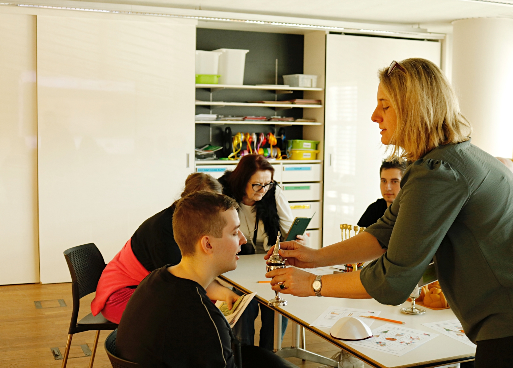 Praktykant ogląda srebrny przedmiot prezentowany przez edukatorkę. Reszta grupy siedzi przy stole.