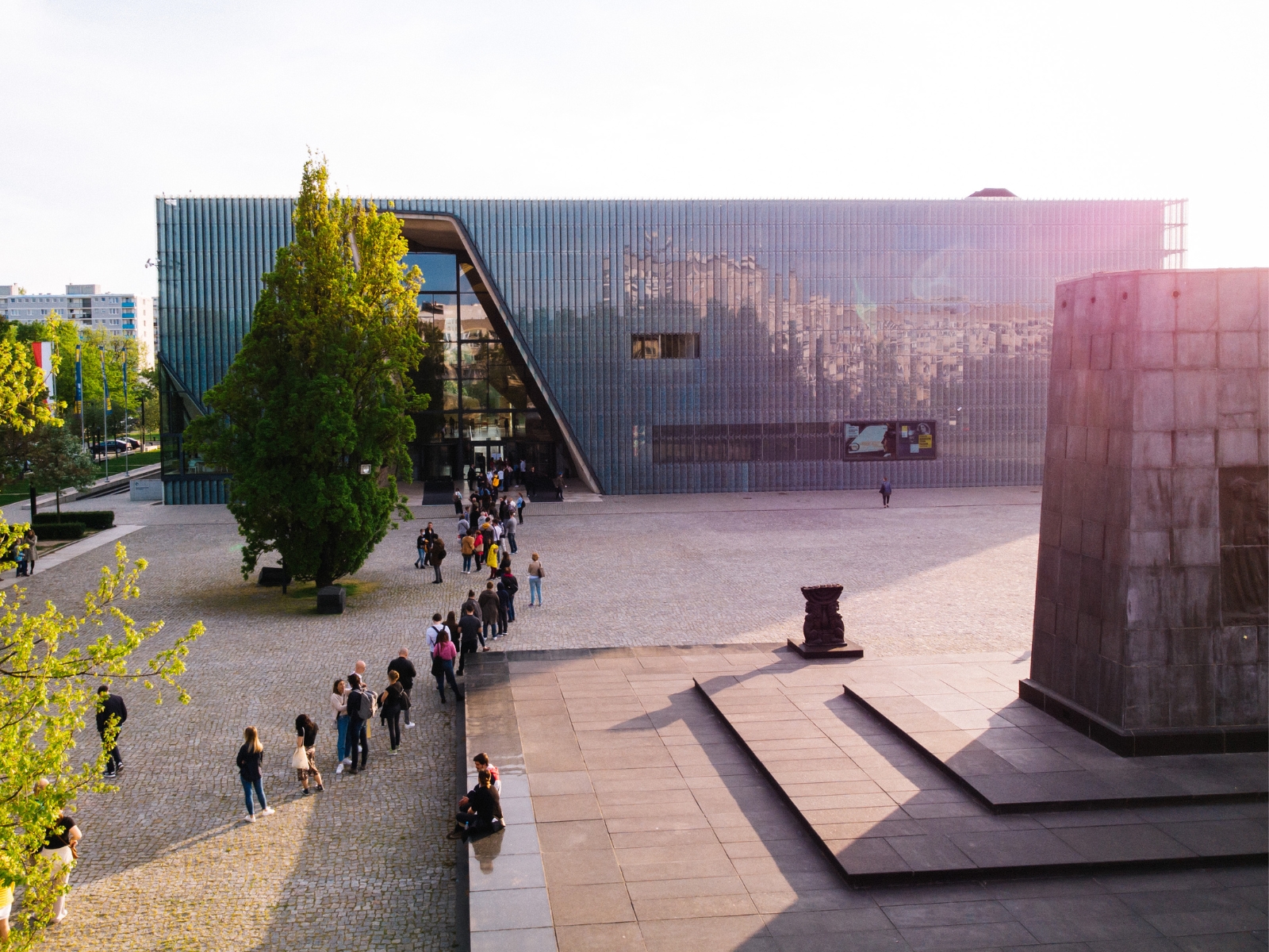Kolejka osób czeka na wejście do budynku Muzeum POLIN. Świeci słońce. Z prawej strony widoczny pomnik Bohaterów Getta.