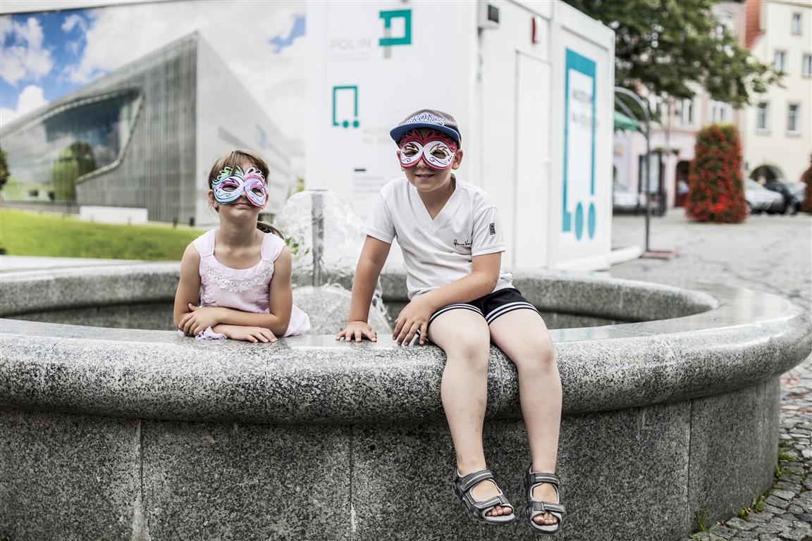 Dwoje dzieci - jedno siedzi na murku wokół fontanny, drugie w środku.
