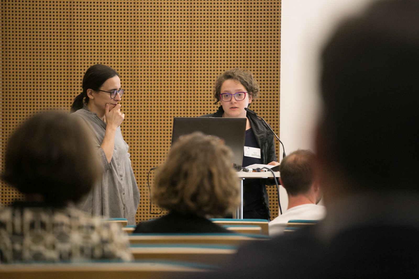 Dwie kobiety przemawiają podczas konferencji. Jedna z nich trzyma dłoń na brodzie.