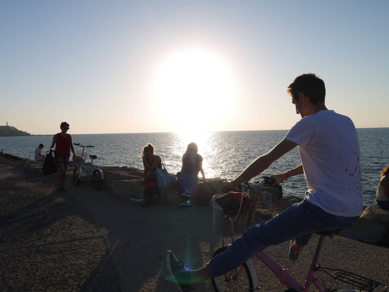 Grupa młodych ludzi na deptaku przy morskim brzegu. Siedzą na murku, jeżdżą na rowerach.  